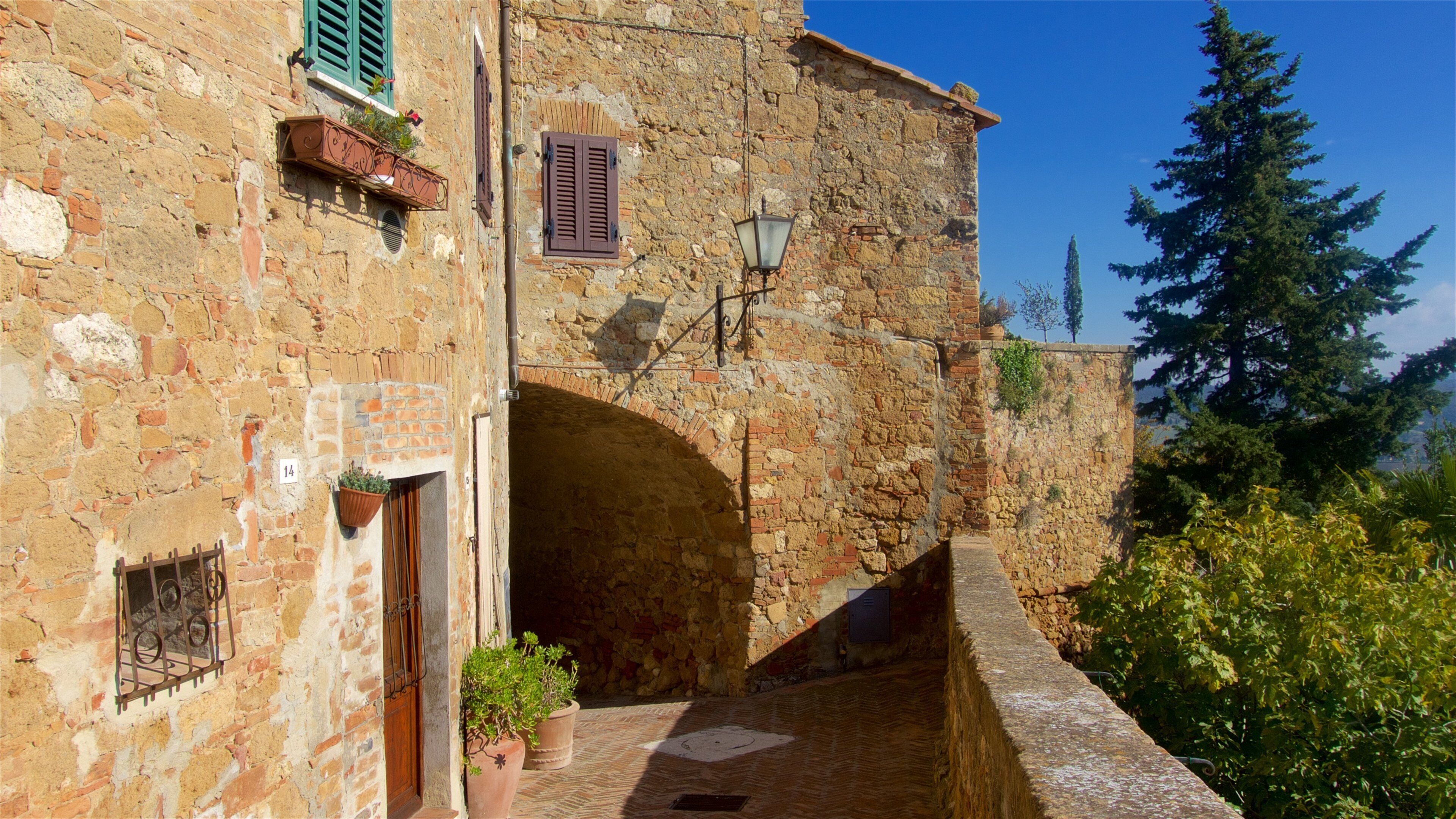 A narrow stone alleyway with a rustic building featuring a wooden door, a metal gate, and potted plants.