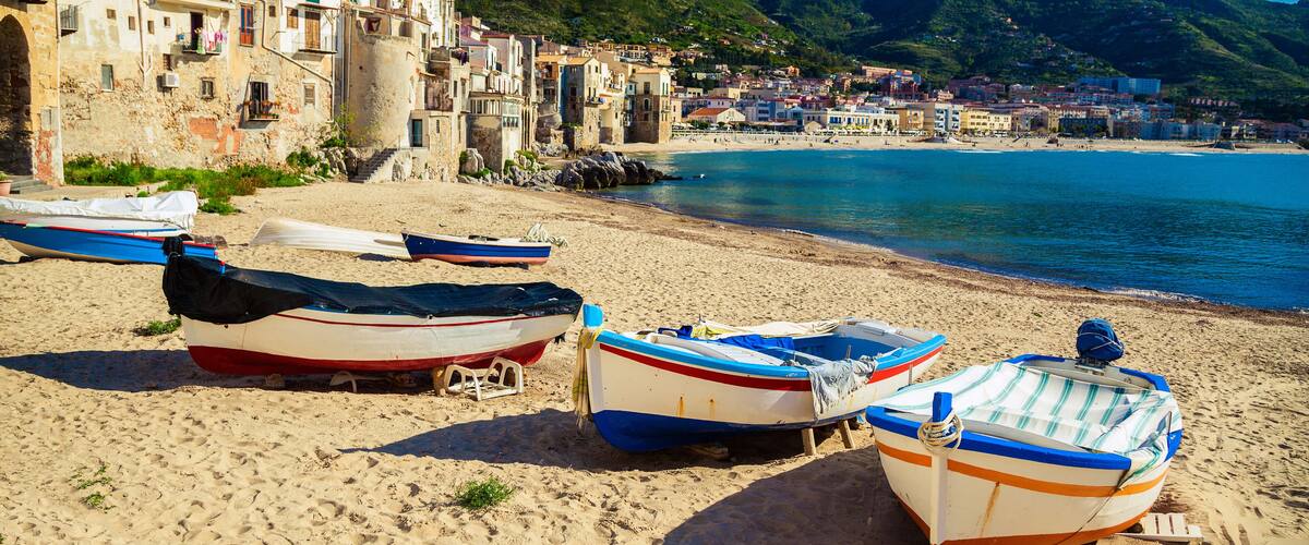 A coastal town with boats on the beach, buildings, and mountains in the background.