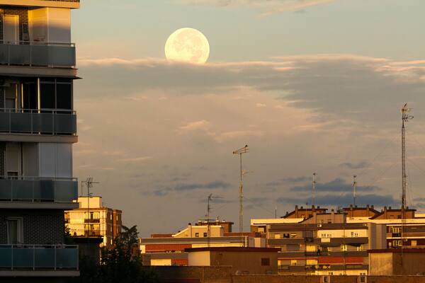 Un paisaje urbano con una luna llena destacada en el cielo, rodeado de edificios con balcones y antenas.