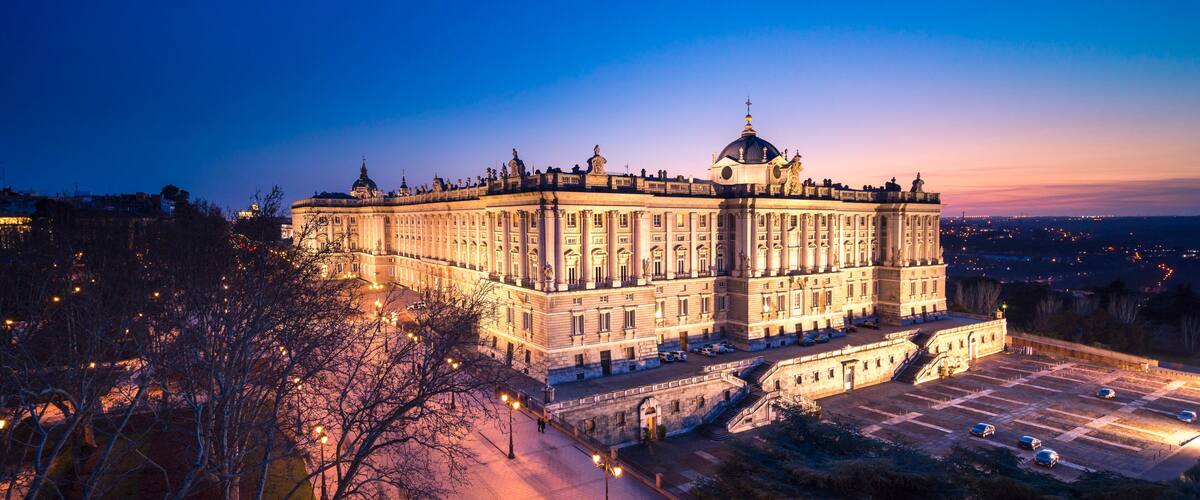 A grand, illuminated building with a dome and multiple towers, surrounded by a spacious courtyard and trees.