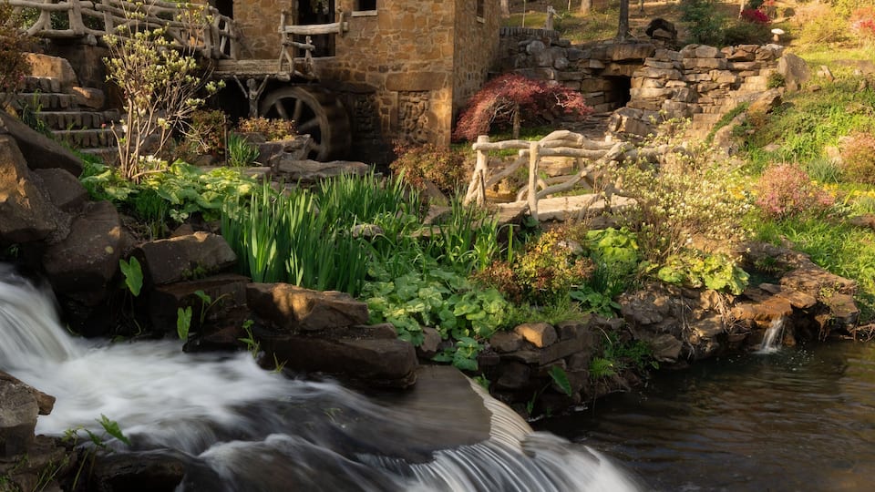 A stone mill house by a flowing stream with surrounding greenery and trees.