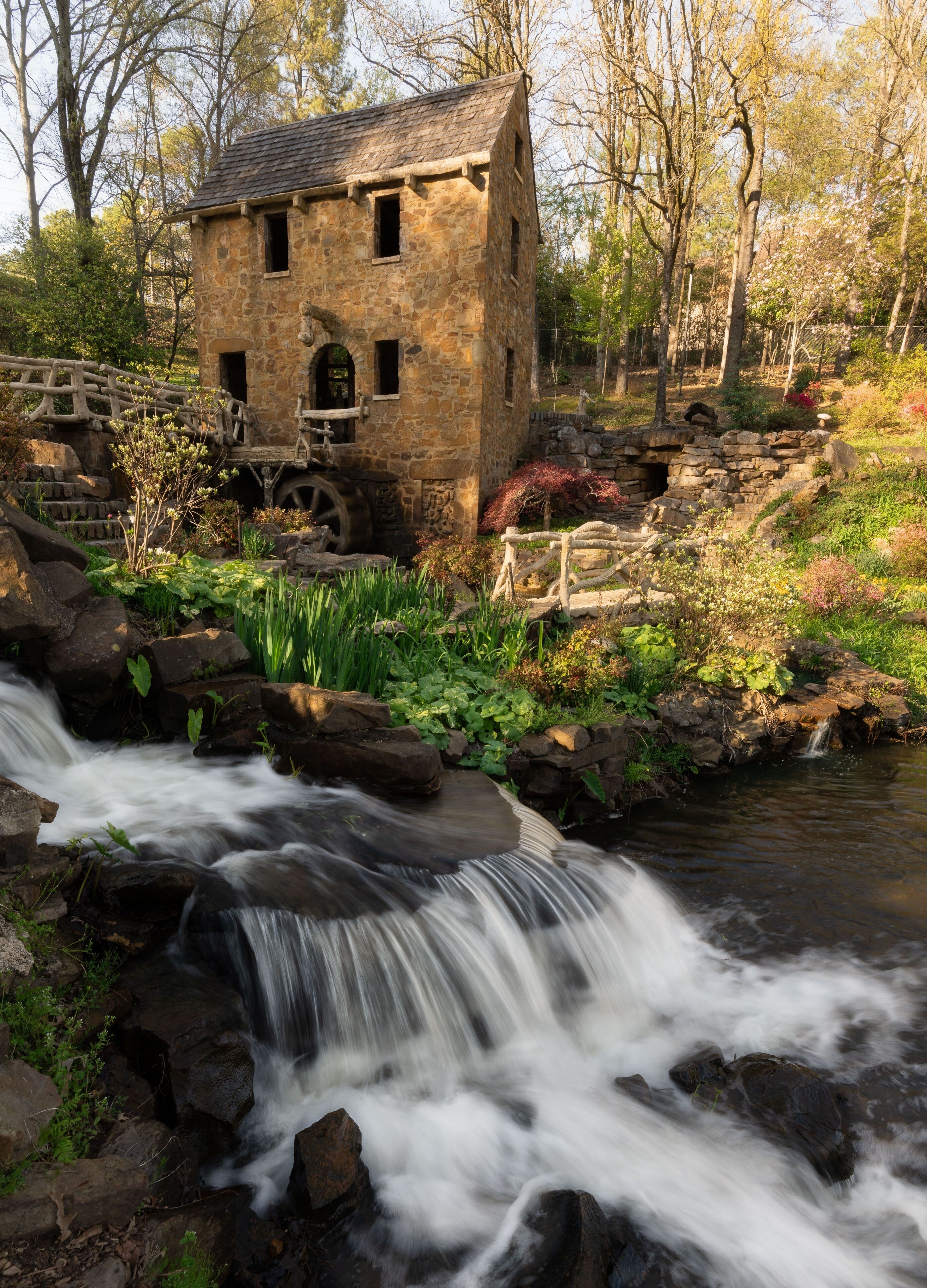 A stone mill house by a flowing stream with surrounding greenery and trees.