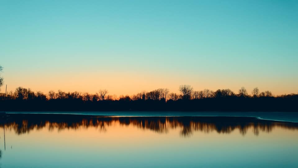 A calm lake at sunset with a silhouette of trees reflecting on the water.