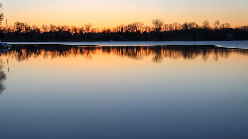 A calm lake at sunset with a reflection of trees and a clear sky.