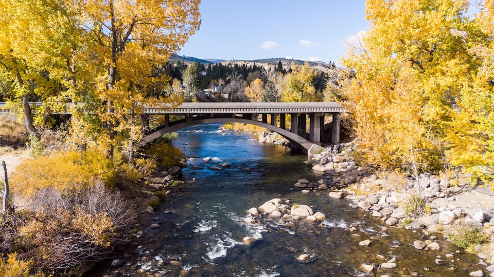 A stone bridge over a clear river surrounded by autumn-colored trees.
