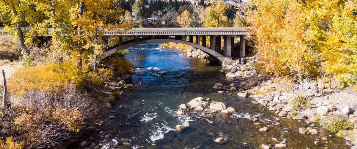 A stone bridge over a clear river surrounded by autumn-colored trees.