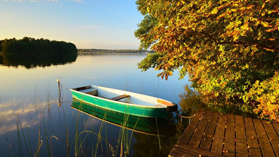 A calm lake with a small green boat tied to a wooden dock, surrounded by trees with autumn leaves.