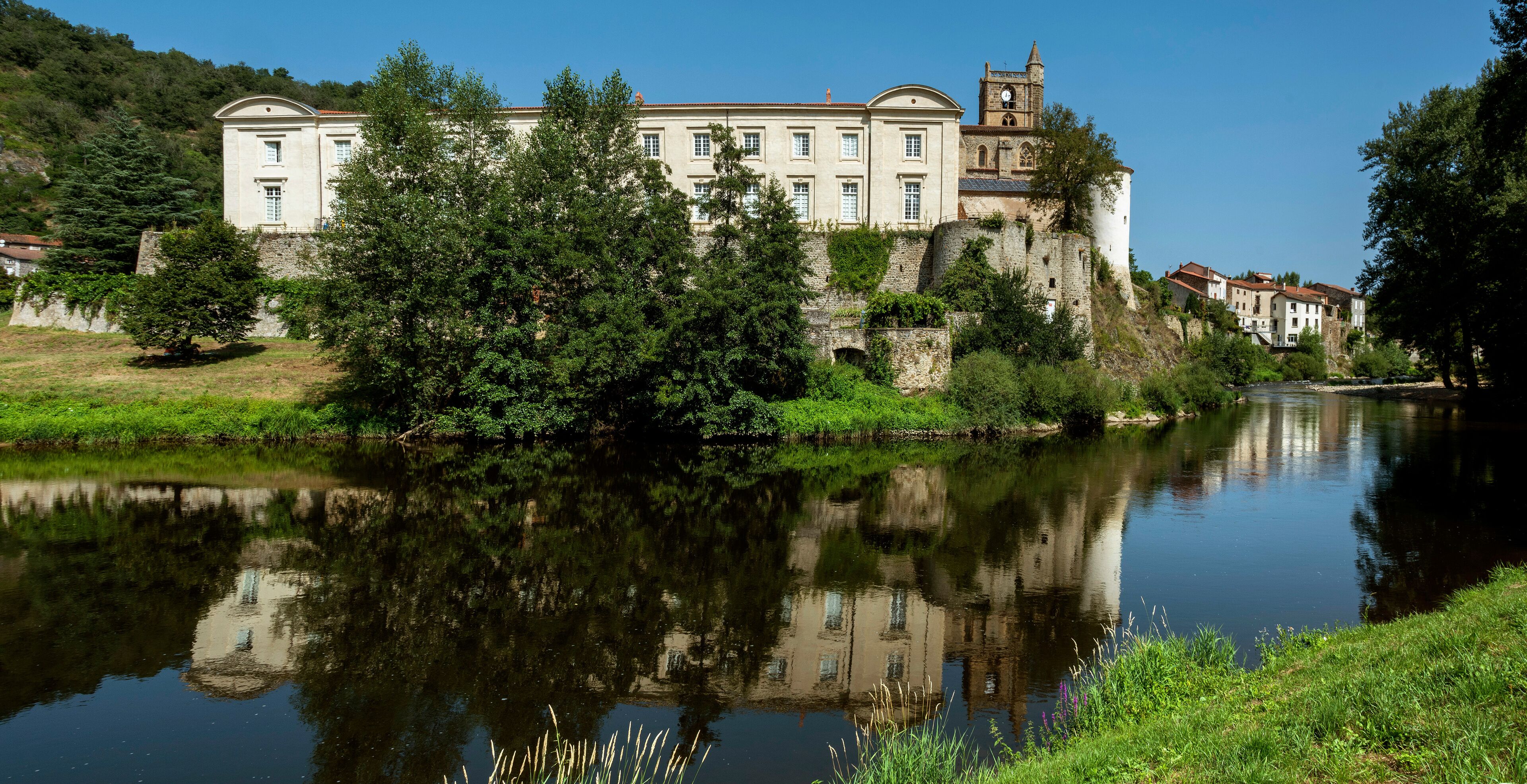 A historic building with a clock tower by a calm river, surrounded by lush greenery and stone walls.