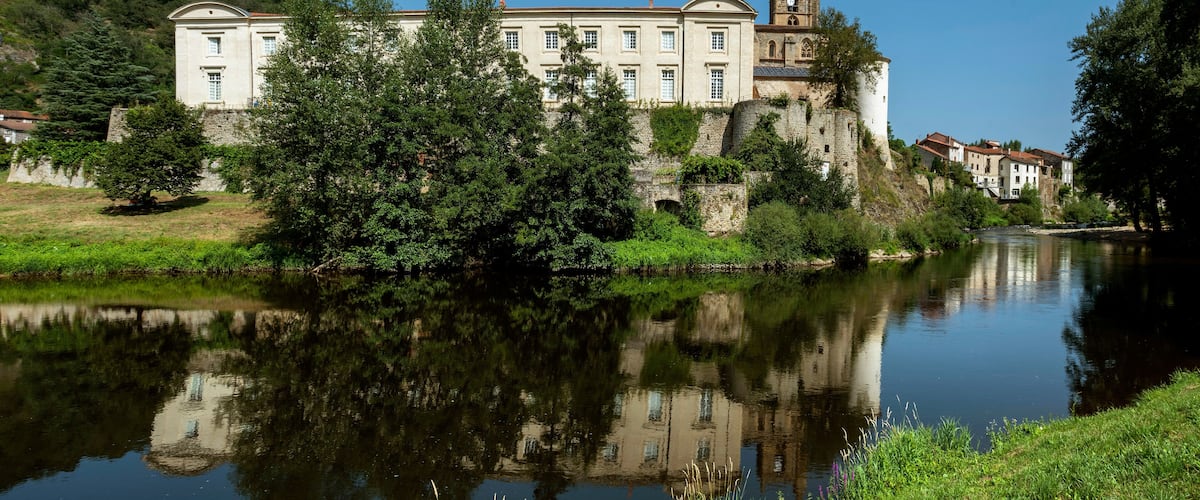A historic building with a clock tower by a calm river, surrounded by lush greenery and stone walls.