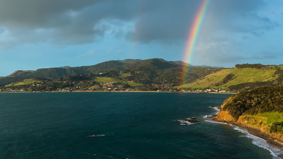 A coastal landscape with a rainbow over the sea, green hills, and a small town by the water.