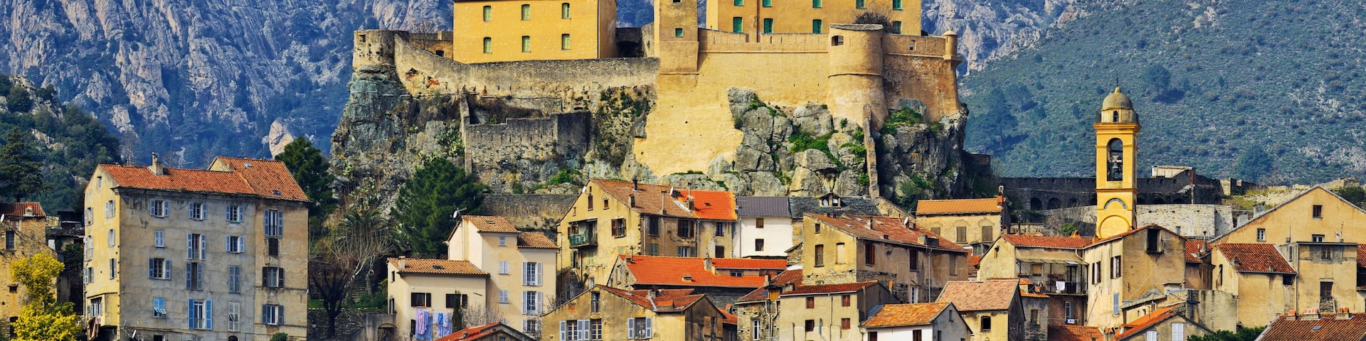A historic fortified town with buildings atop a cliff, surrounded by rugged mountains.
