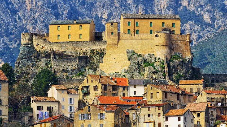 A historic fortified town with buildings atop a cliff, surrounded by rugged mountains.