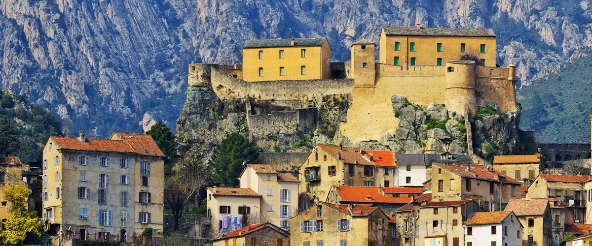 A historic fortified town with buildings atop a cliff, surrounded by rugged mountains.
