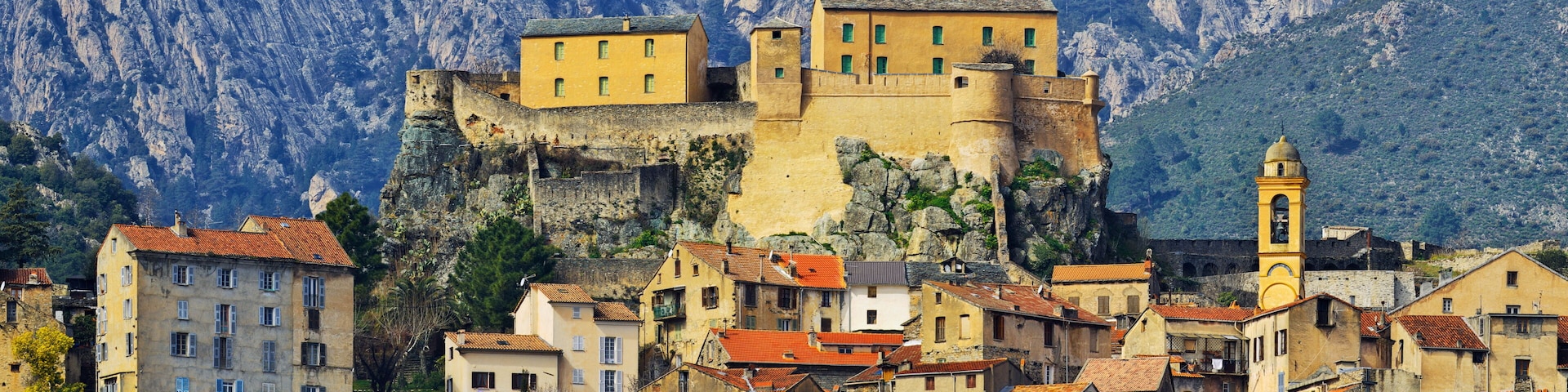 A historic fortified town with buildings atop a cliff, surrounded by rugged mountains.