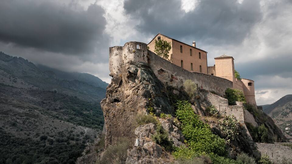A medieval castle perched on a rocky cliff with a mountainous backdrop.