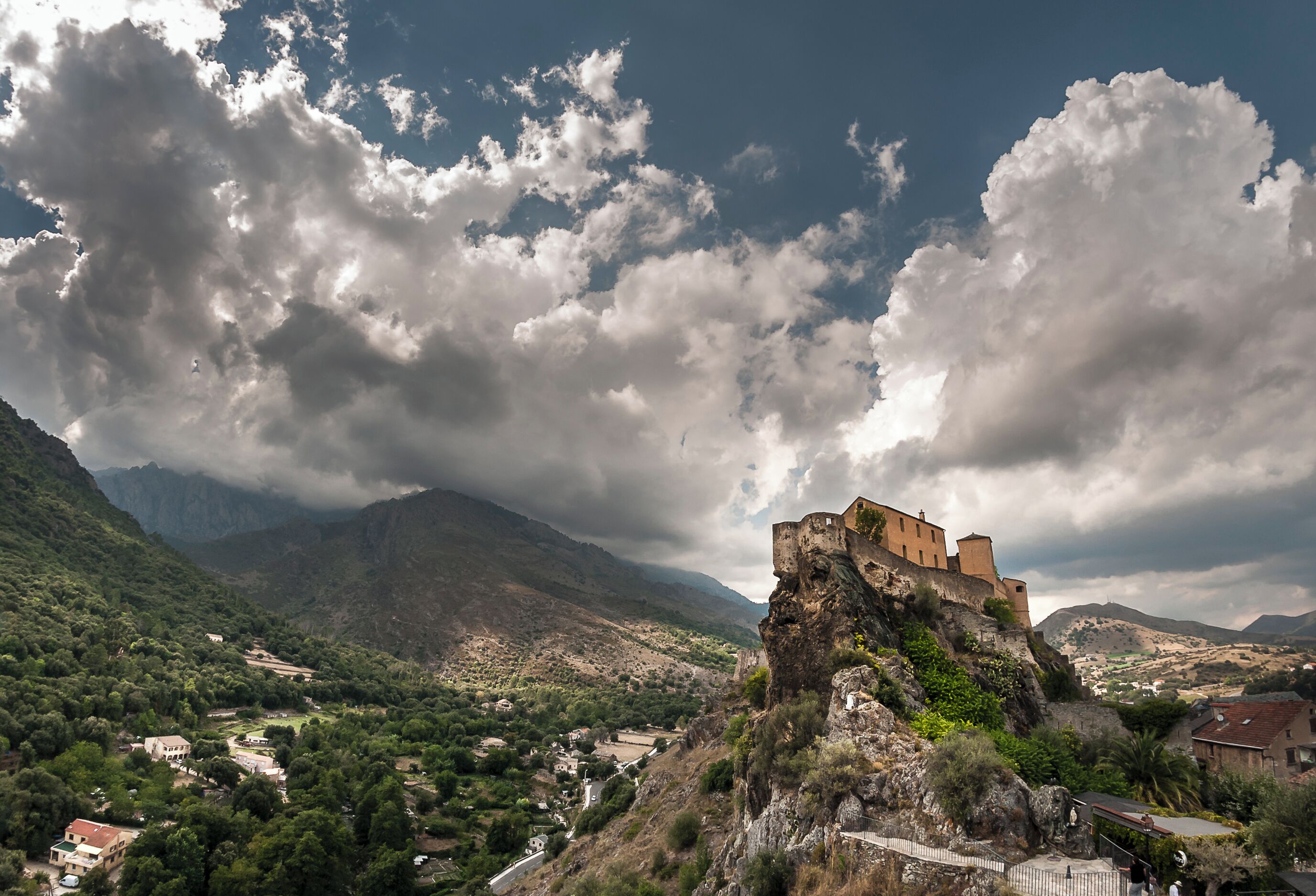 A historic castle on a rocky cliff overlooking a valley with mountains in the background.