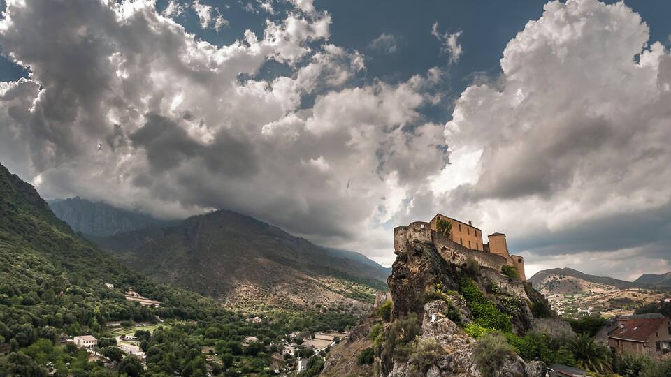 A historic castle on a rocky cliff overlooking a valley with mountains in the background.