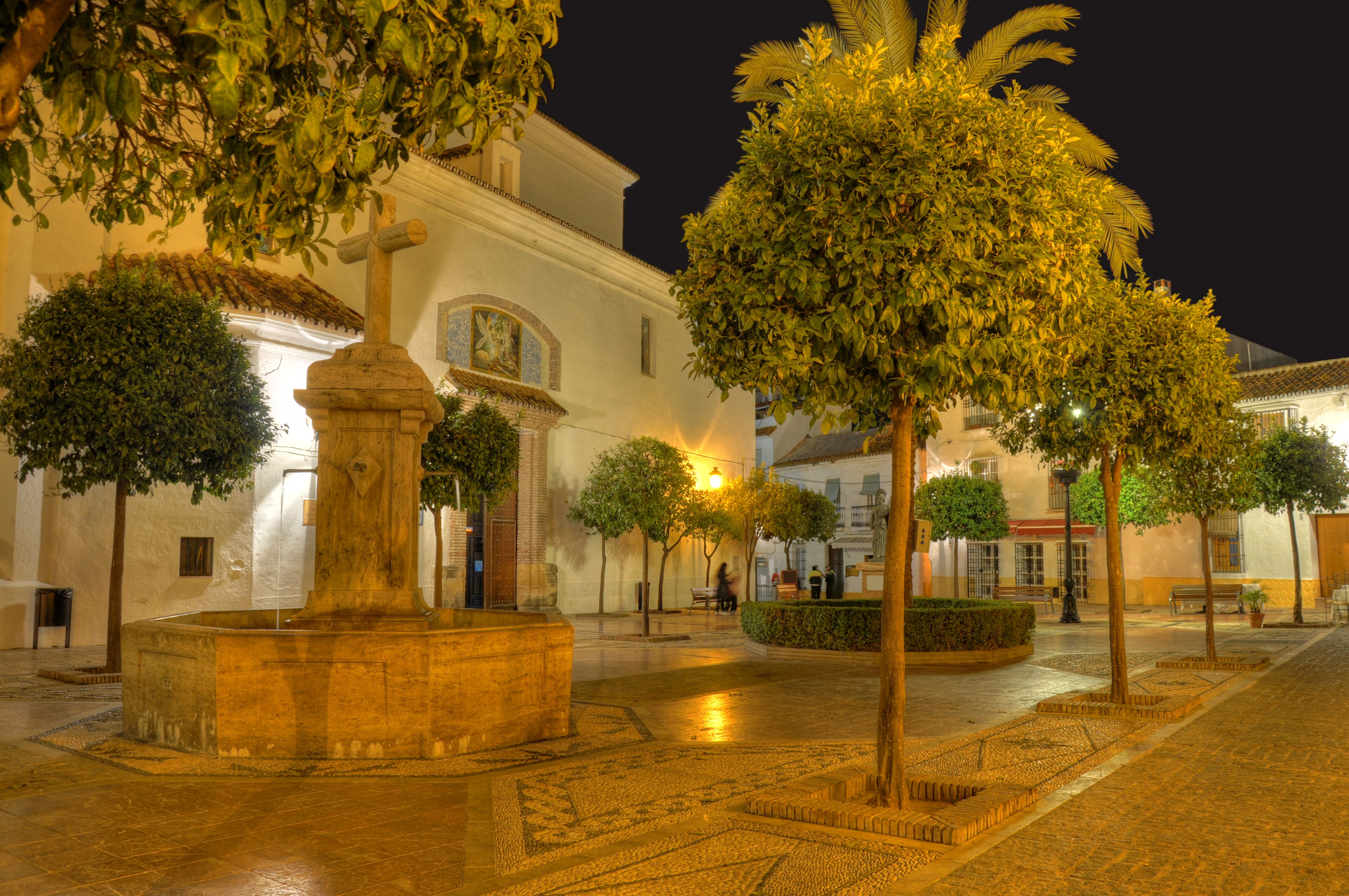 A historic plaza with a fountain, trees, and a white building with a tiled roof.