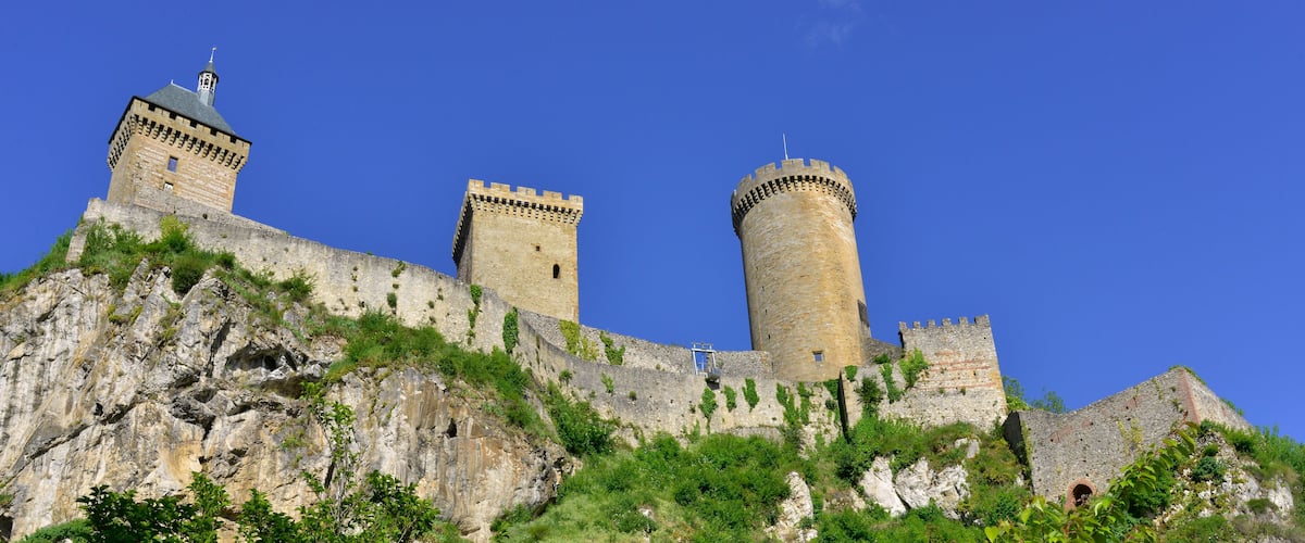 A medieval castle with two prominent towers and a large rock base, surrounded by greenery.