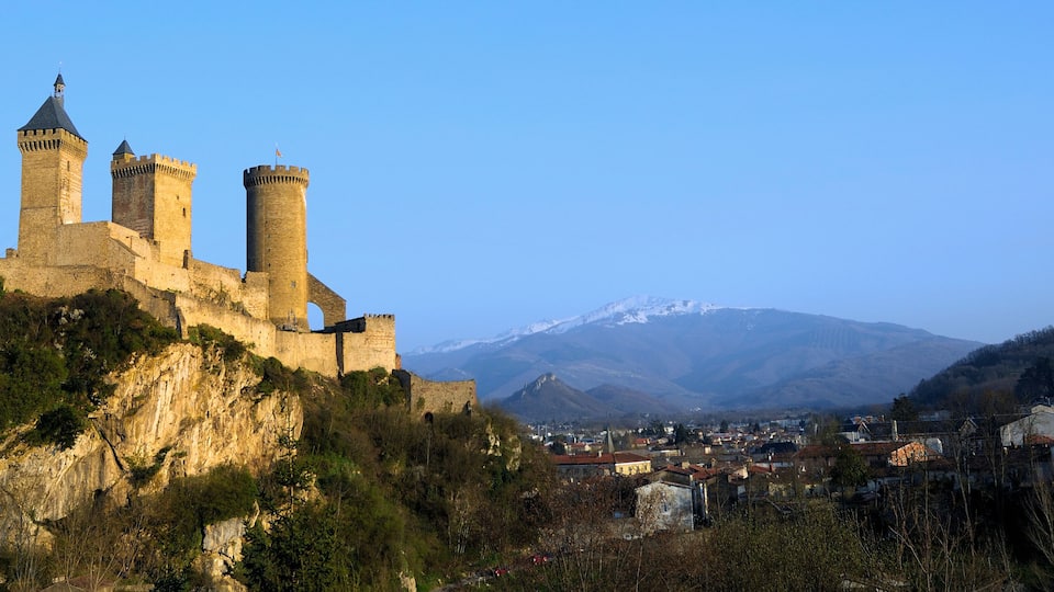 A historic castle with two towers on a hill, overlooking a town and a mountain range.