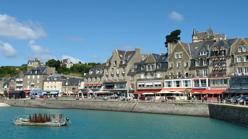 A waterfront town with historic buildings, a boat docked at the pier, and people walking along the promenade.