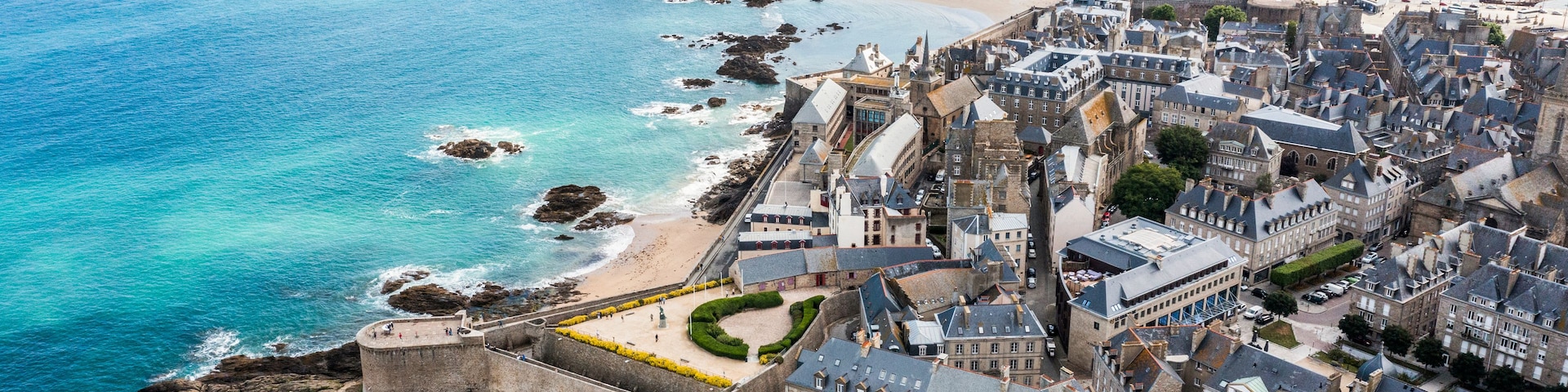 Un pueblo costero con una fortificación histórica, una playa de arena y un mar azul cristalino.