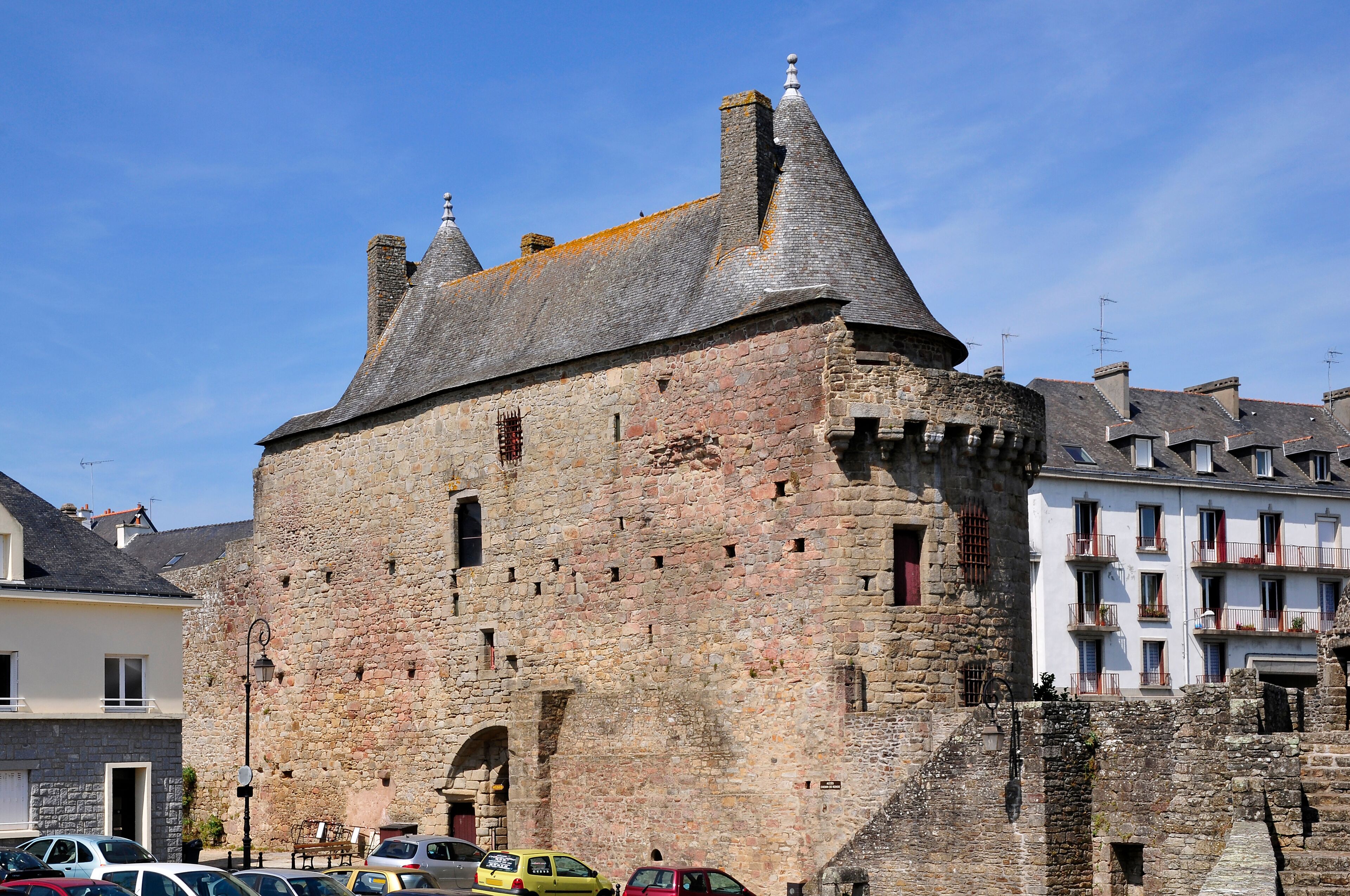 A historic stone castle with a conical roof, surrounded by modern buildings and parked cars.