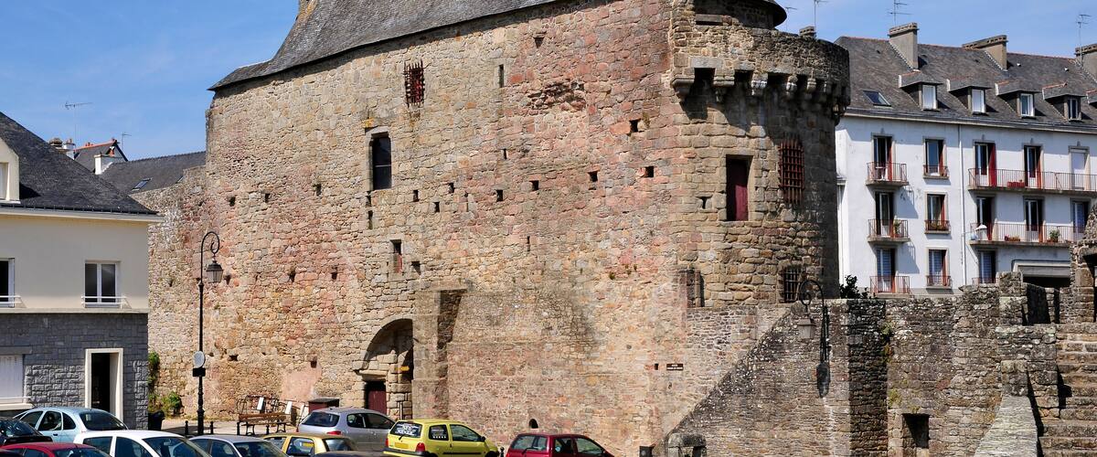 A historic stone castle with a conical roof, surrounded by modern buildings and parked cars.