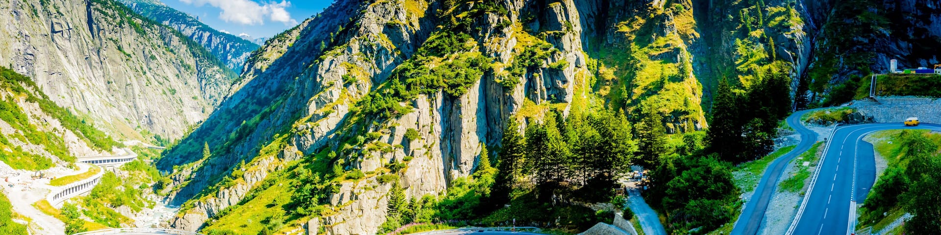 A winding mountain road with a guardrail, surrounded by steep cliffs and lush greenery.