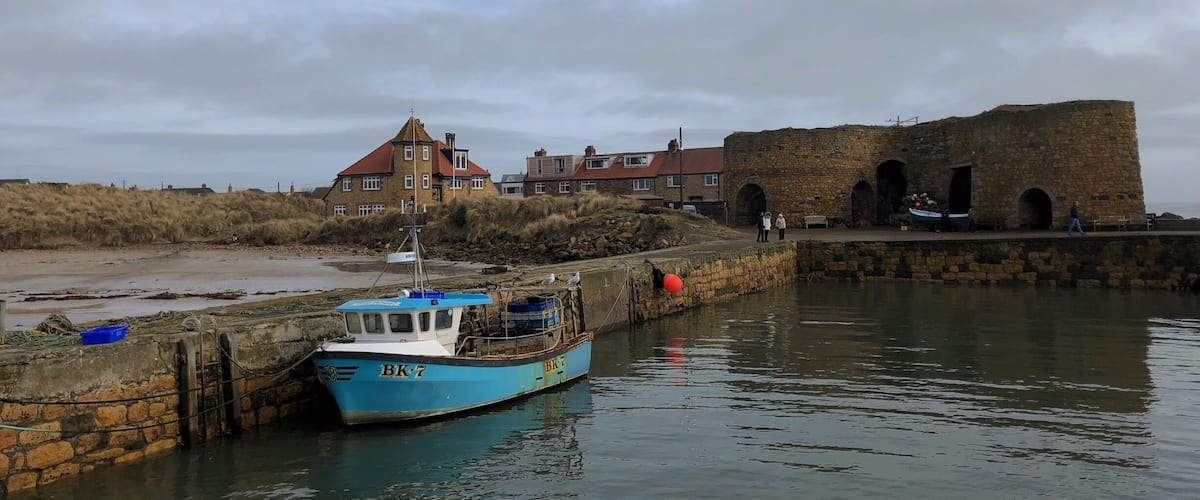 A coastal scene with a blue boat docked at a stone pier, a historic stone building, and a sandy beach with dunes in the background.