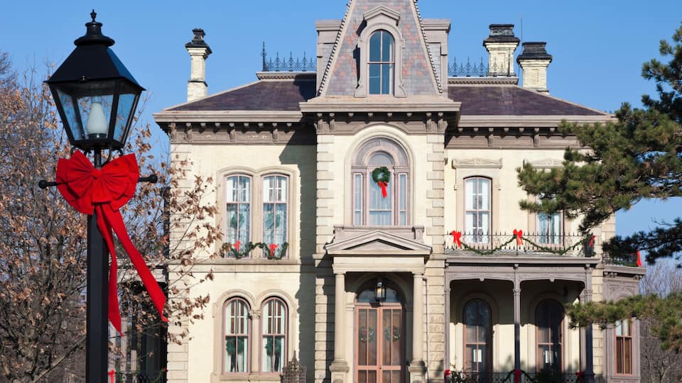 A large, historic house with a prominent front entrance, a red bow on a street lamp, and a clear blue sky.
