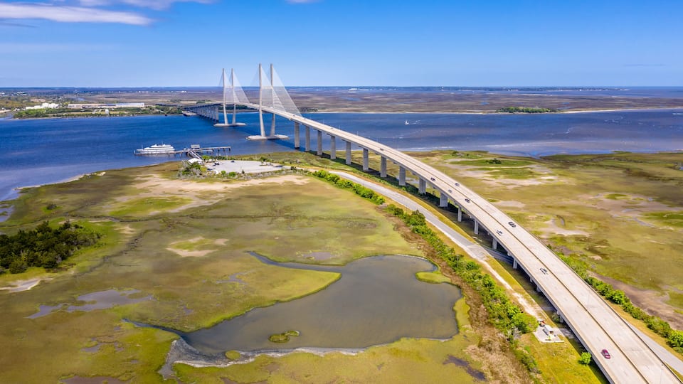 A long bridge spanning a wide river with surrounding wetlands and a distant shoreline.