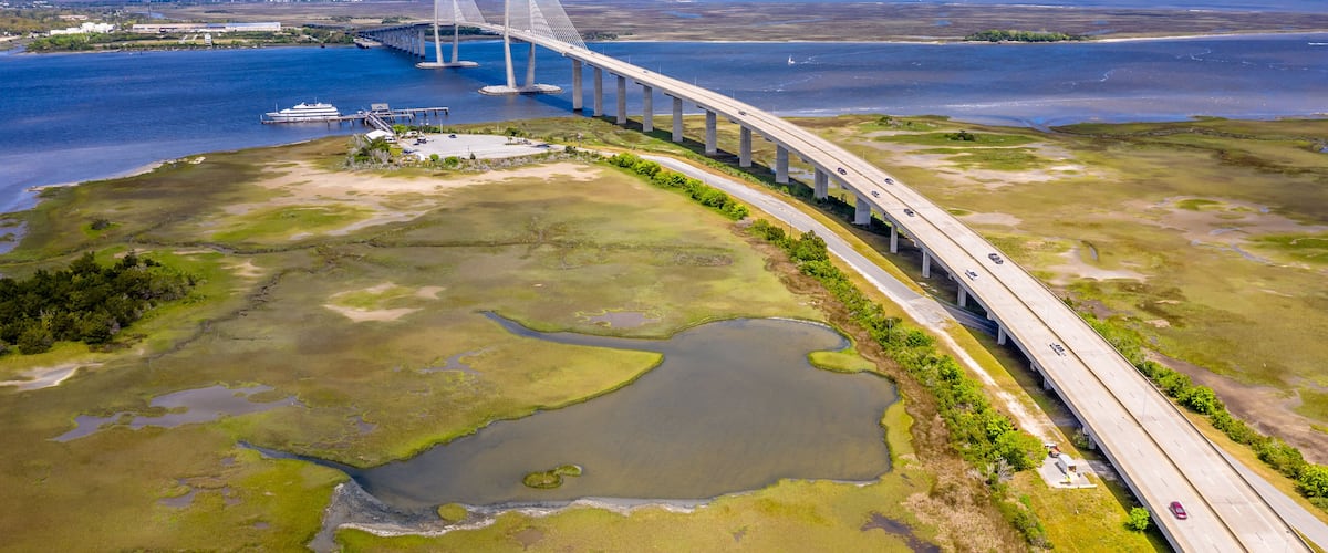 A long bridge spanning a wide river with surrounding wetlands and a distant shoreline.