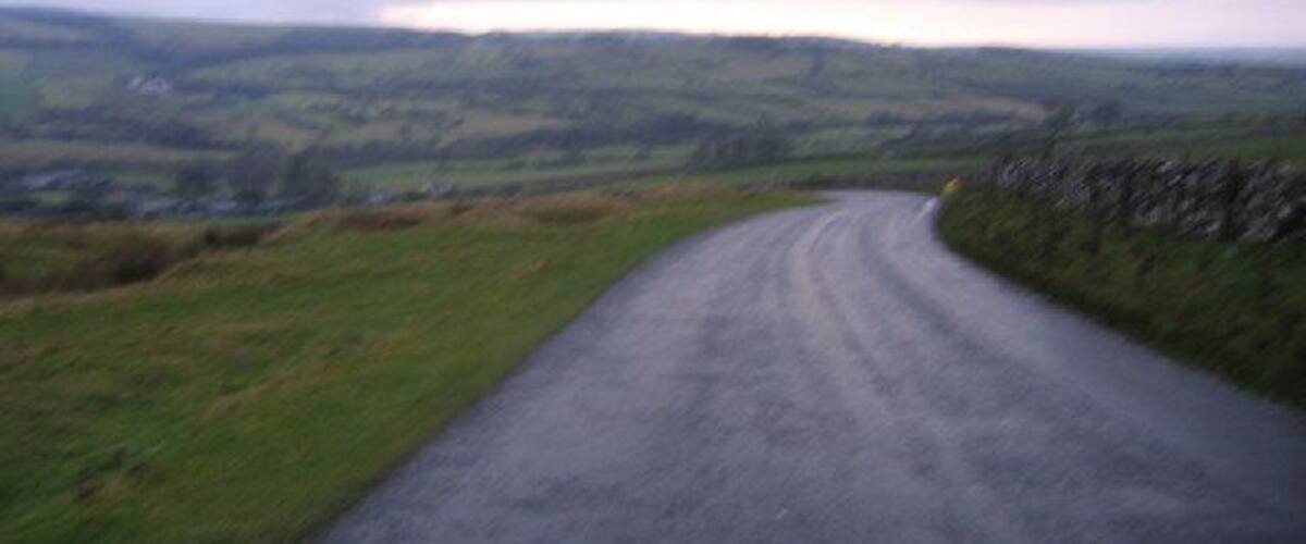 A curving country road with stone walls on one side and grassy fields on the other.