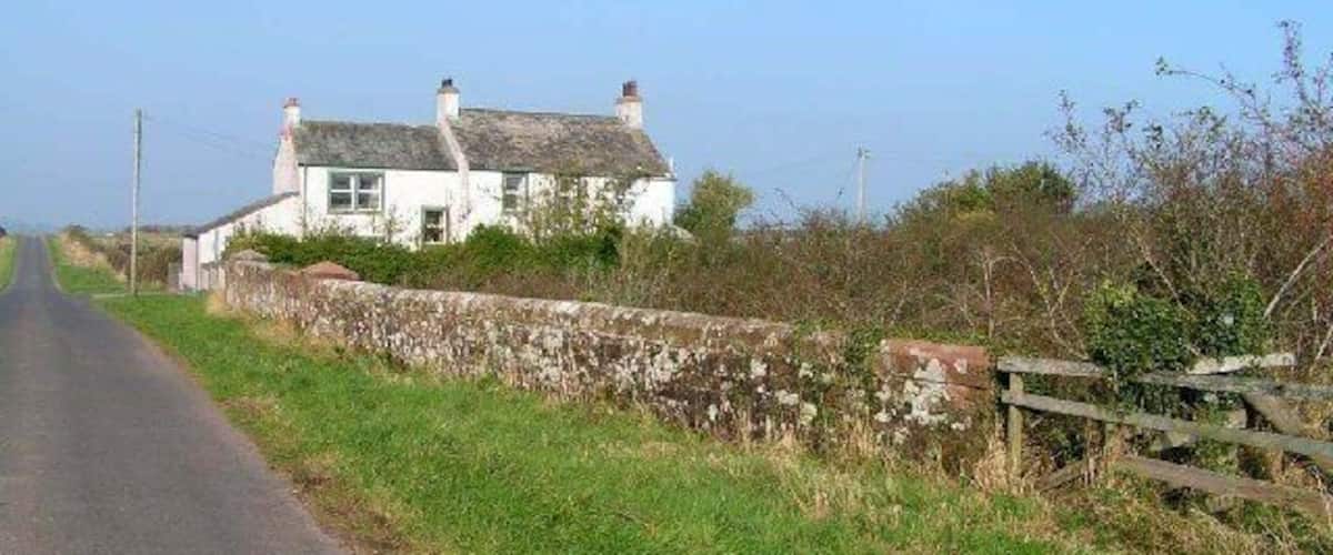 A rural road with a white house on the left, stone wall, and wooden fence.