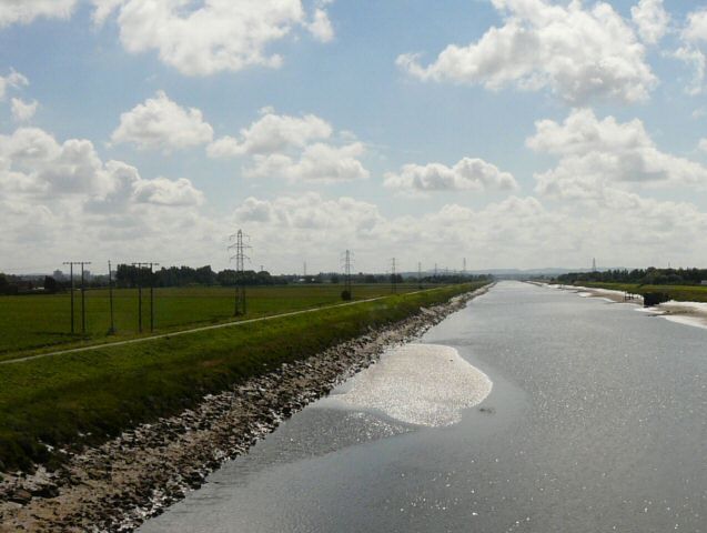 A river with a rocky bank, green fields, and power lines in the distance.