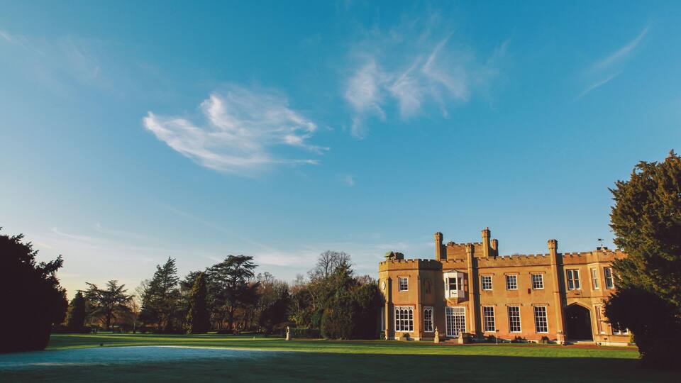 A large, historic building with a symmetrical facade, surrounded by a well-manicured lawn and dense trees.