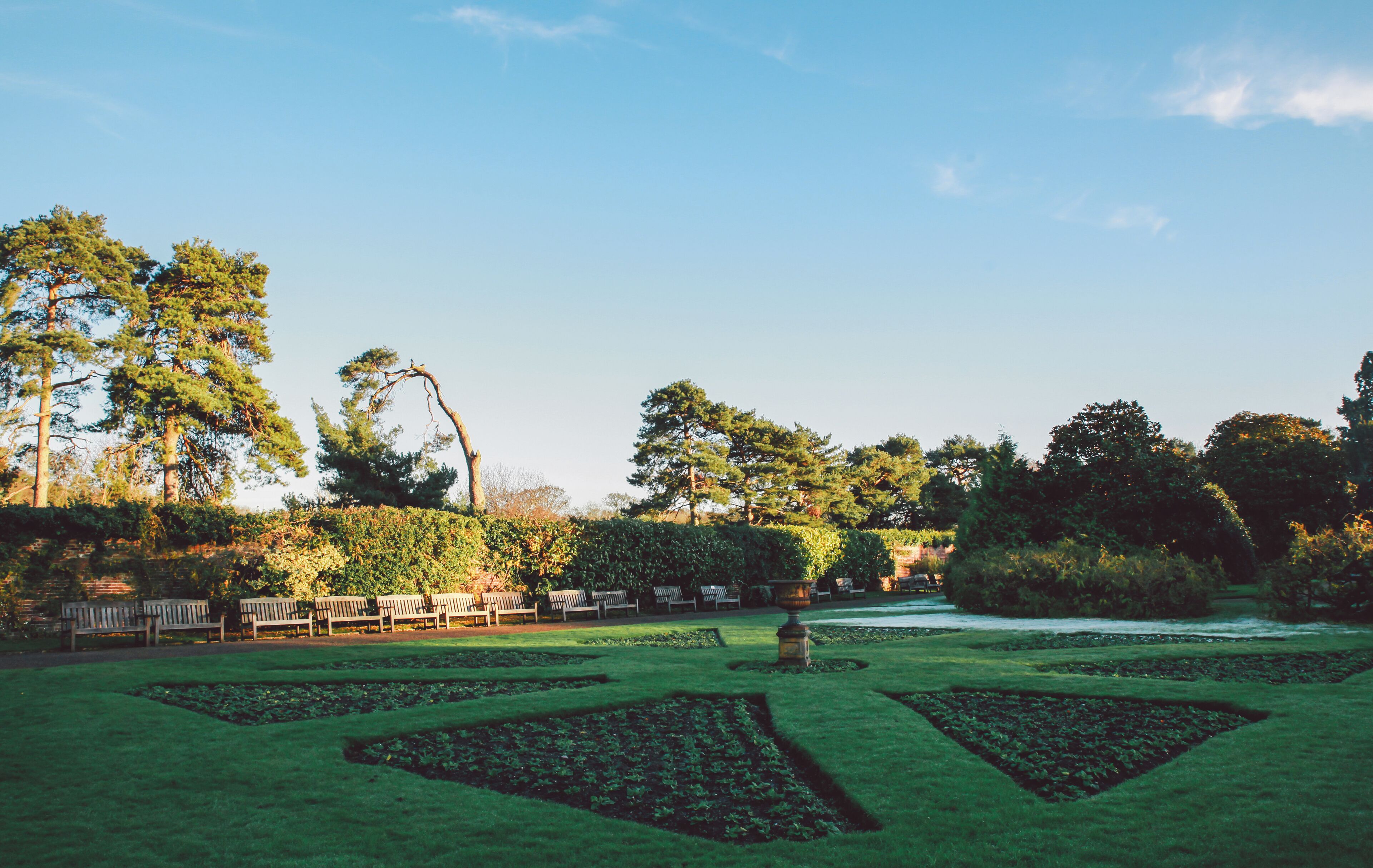 A well-manicured garden with geometric flower beds, a row of wooden benches, and tall trees under a clear sky.