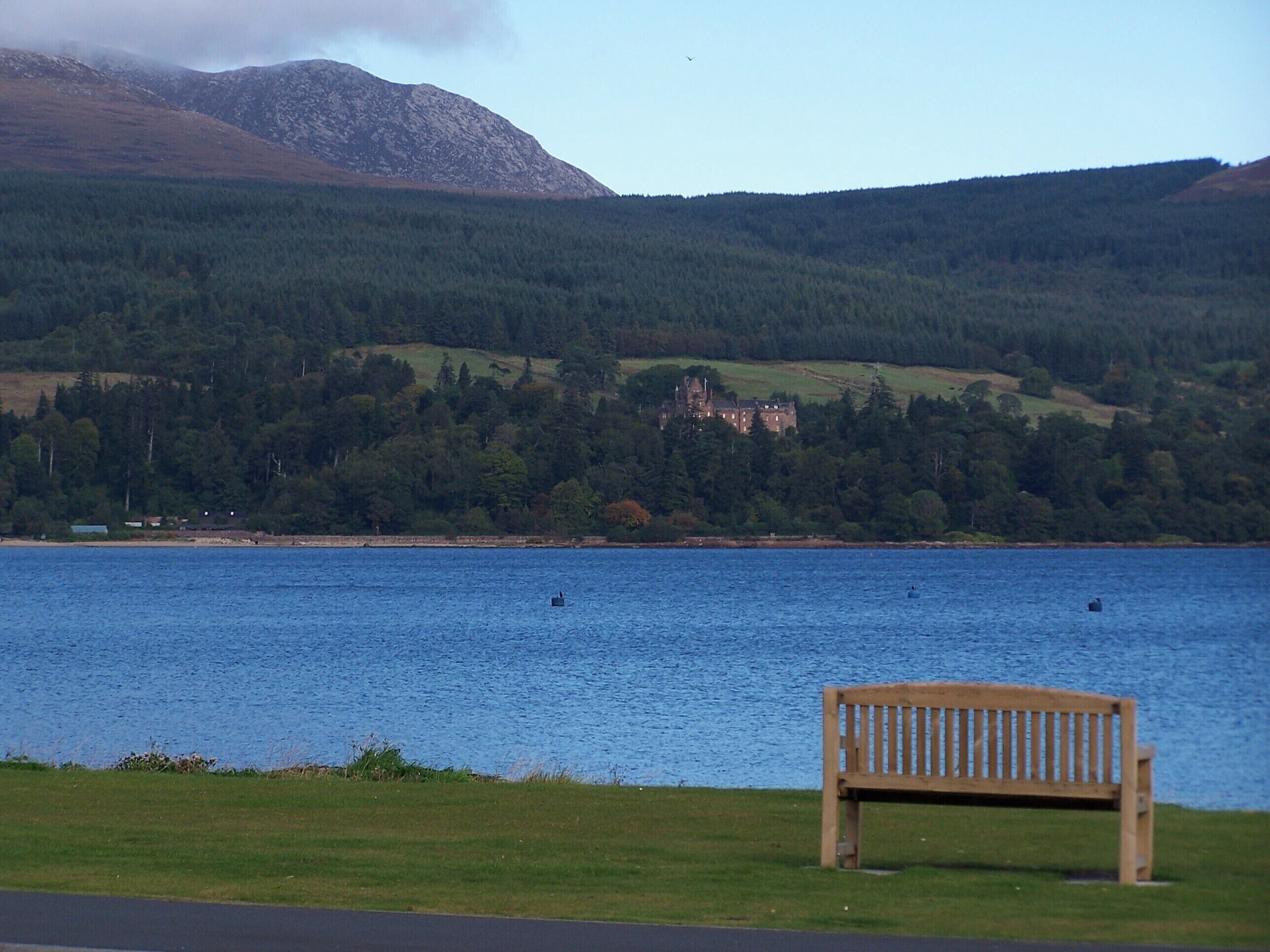 A lake with a wooden bench in the foreground, a castle on a hill in the background, and mountains in the distance.