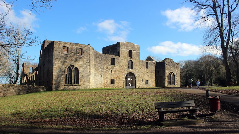 Un bâtiment historique en pierre, doté de fenêtres en arc de cercle et d’une tour centrale, entouré d’une pelouse et d’arbres.