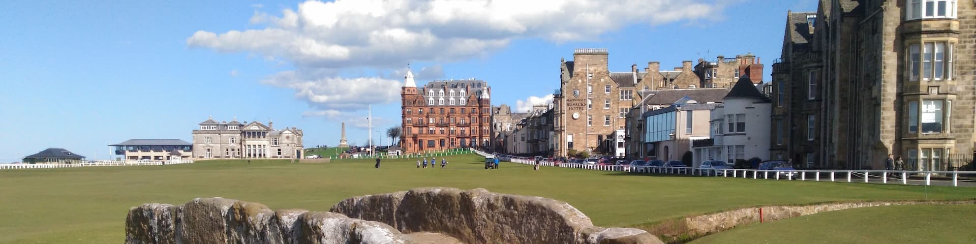 A stone bridge over a grassy field with historic buildings in the background.