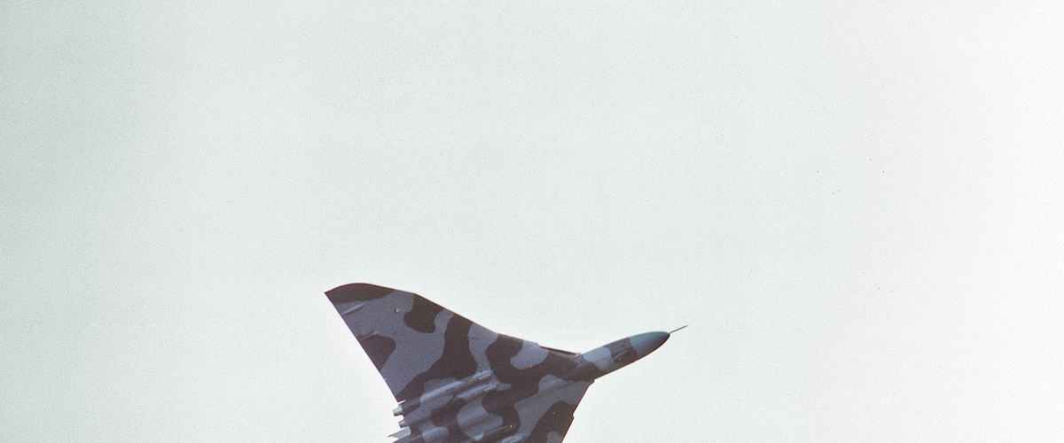 A military aircraft with camouflage pattern flying through a cloudy sky.