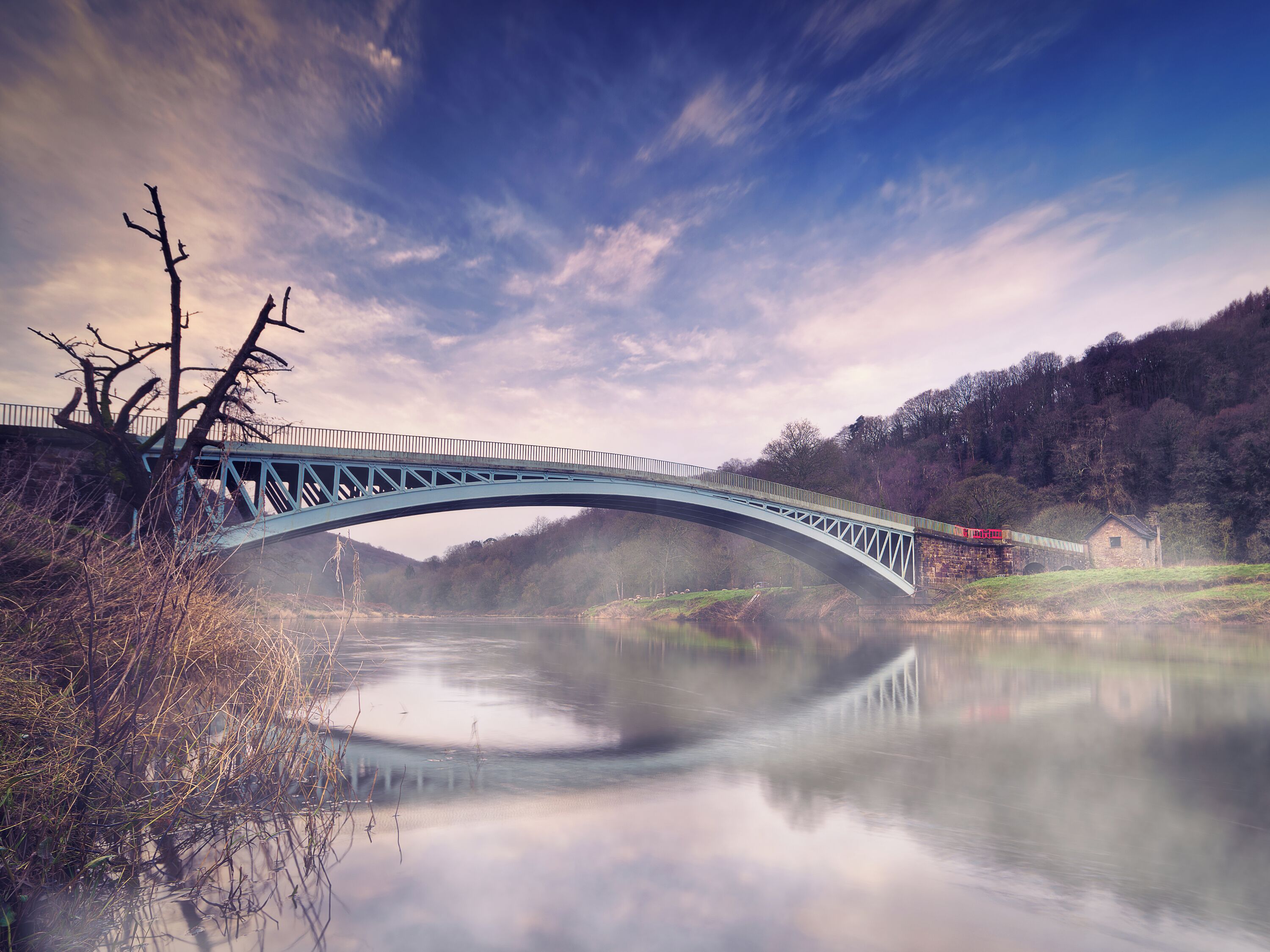 A blue bridge over a calm river, surrounded by trees and a misty atmosphere.