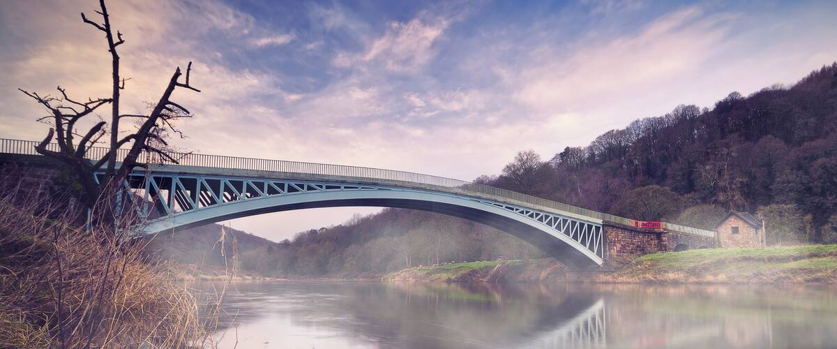 A blue bridge over a calm river, surrounded by trees and a misty atmosphere.