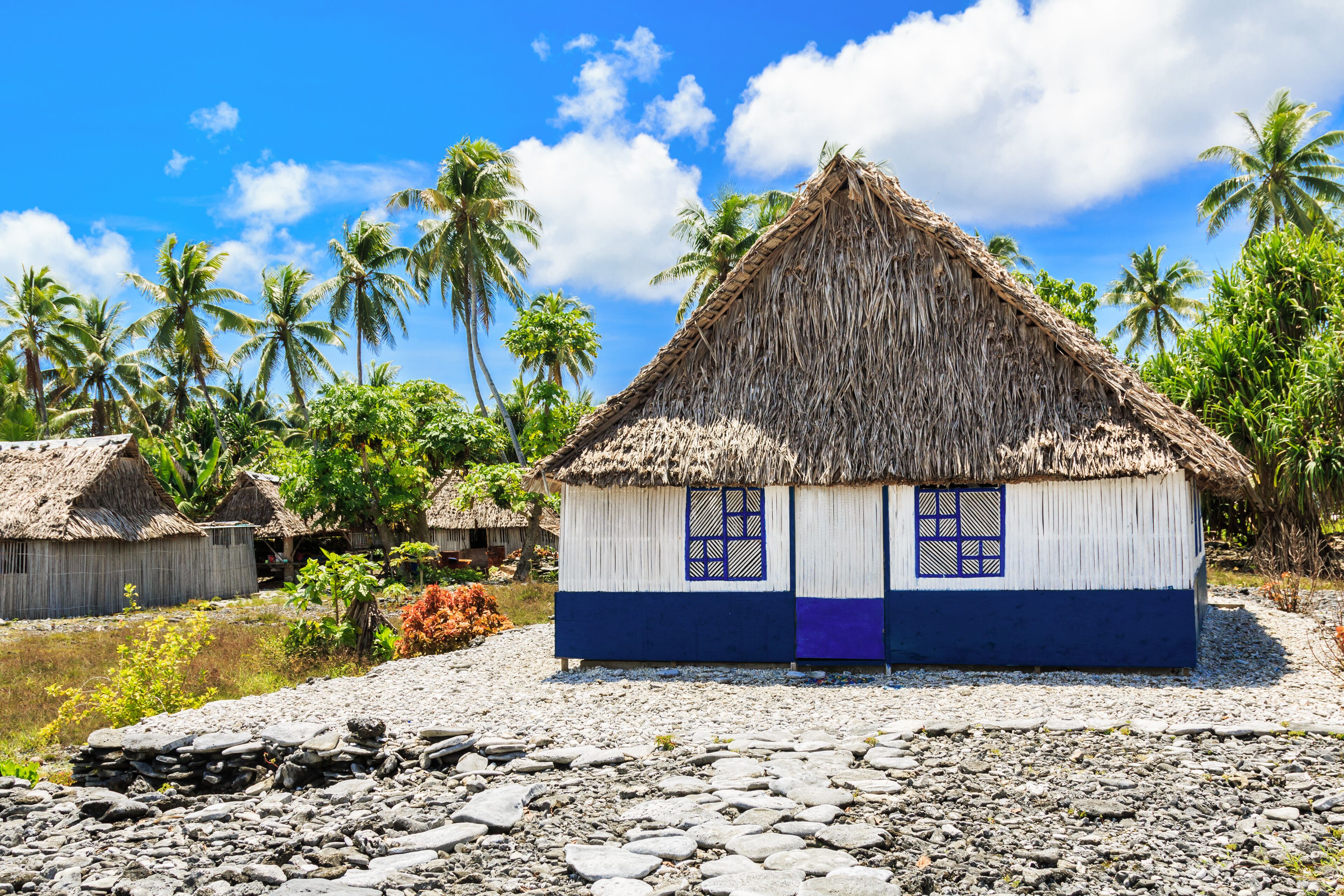 A thatched-roof house with blue and white walls, surrounded by palm trees and a clear blue sky.