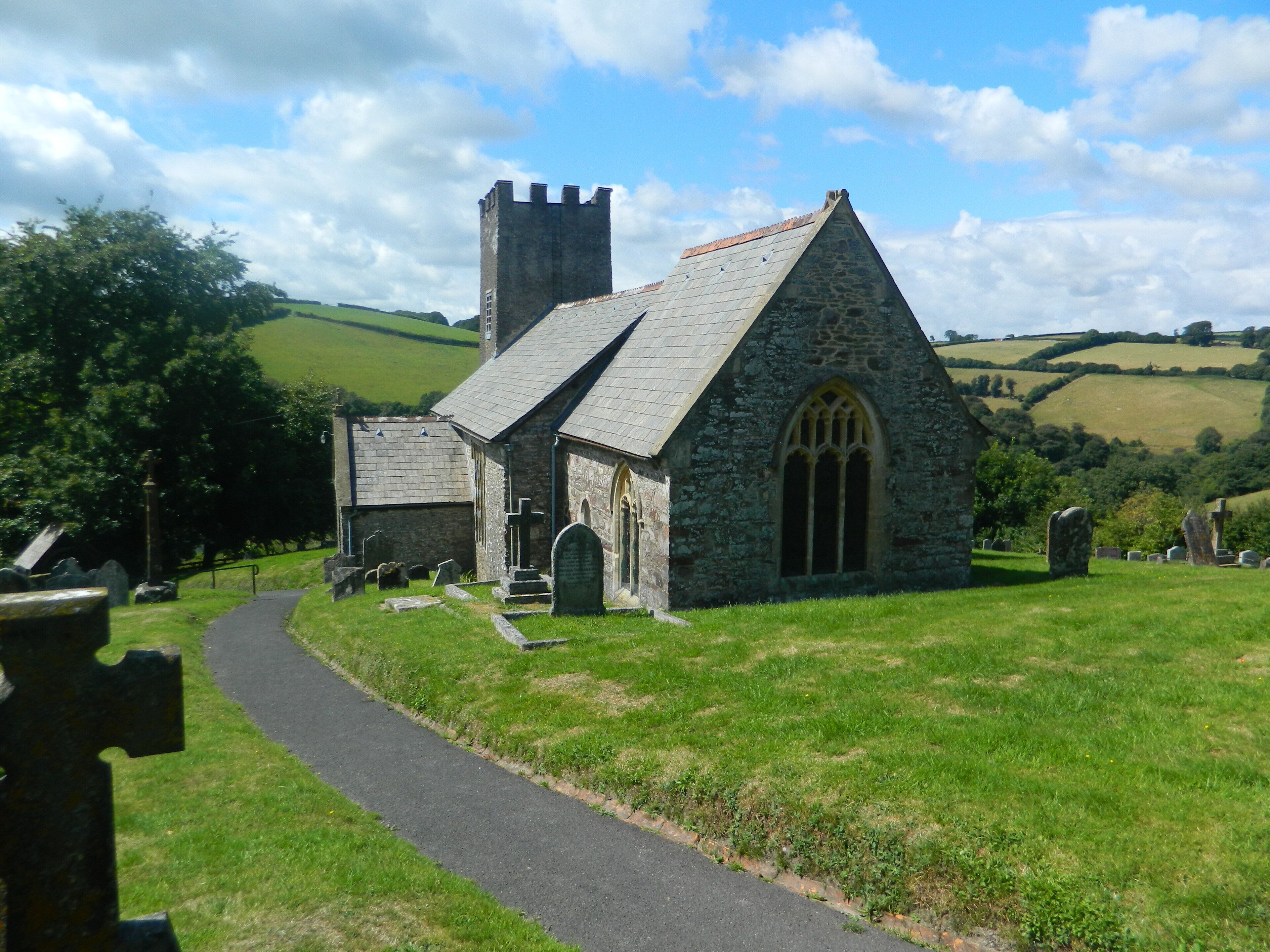 A stone church with a bell tower, surrounded by a cemetery and rolling hills.