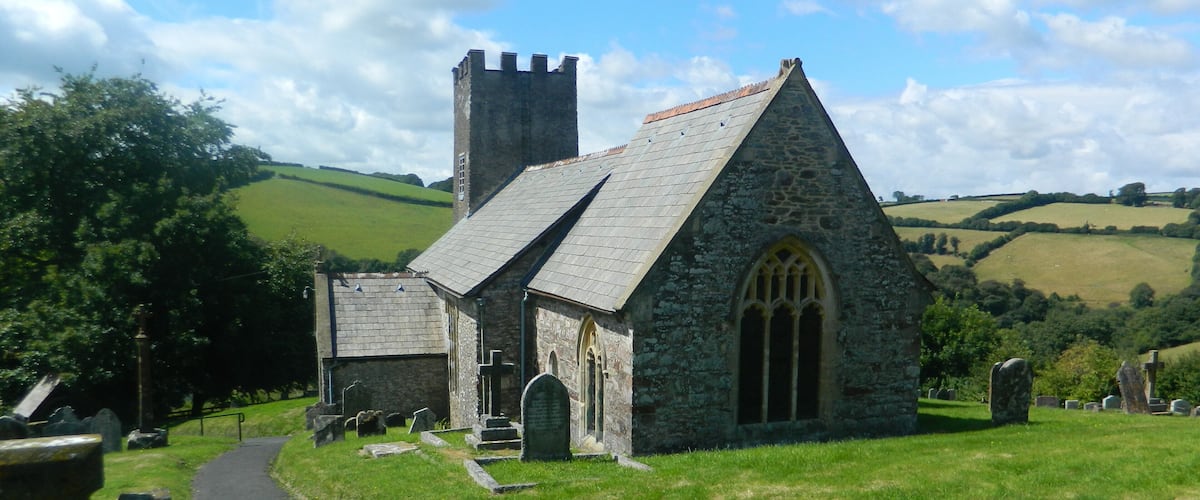 A stone church with a bell tower, surrounded by a cemetery and rolling hills.
