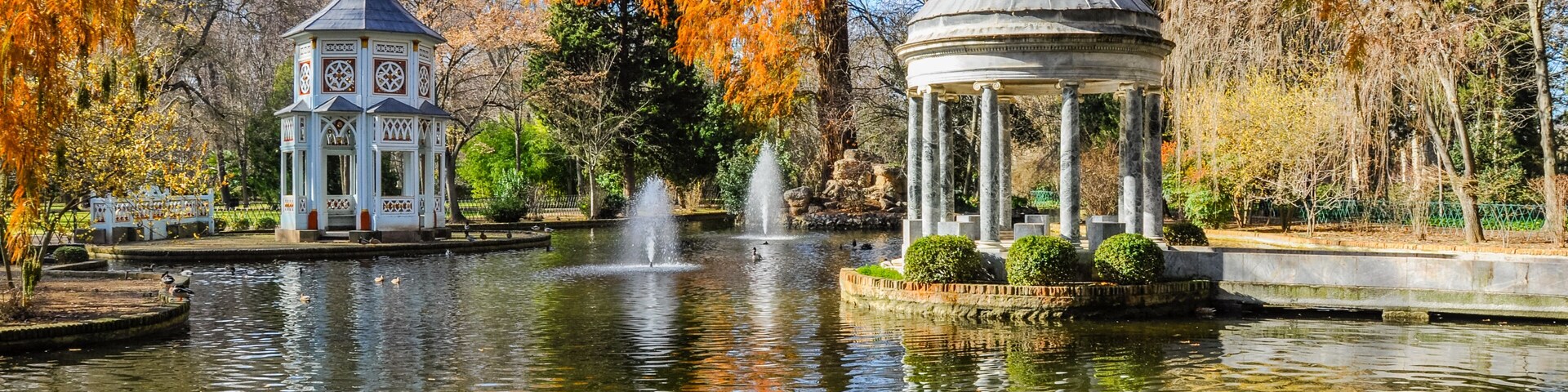 A tranquil park with a pond, a gazebo, and a pavilion with a fountain.