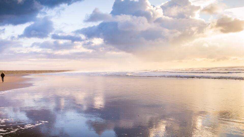 A beach with two people walking, waves crashing, and a cloudy sky.