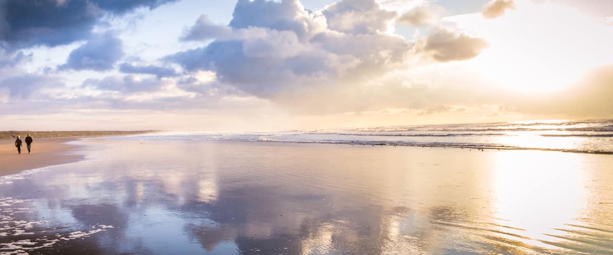 A beach with two people walking, waves crashing, and a cloudy sky.